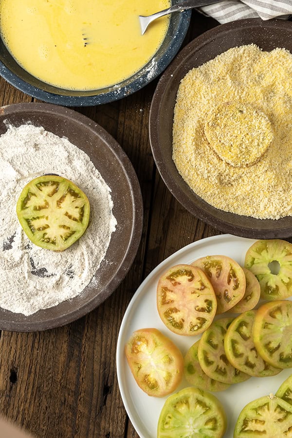 Sliced green tomatoes on a white plate with three stage breading ingredients in enamel pie plates