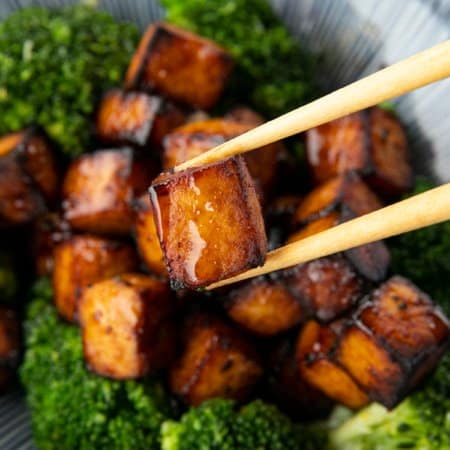 Crispy, golden brown cubes of air fryer tofu served with a drizzle of honey and broccoli on a blue and white striped oblong bowl.