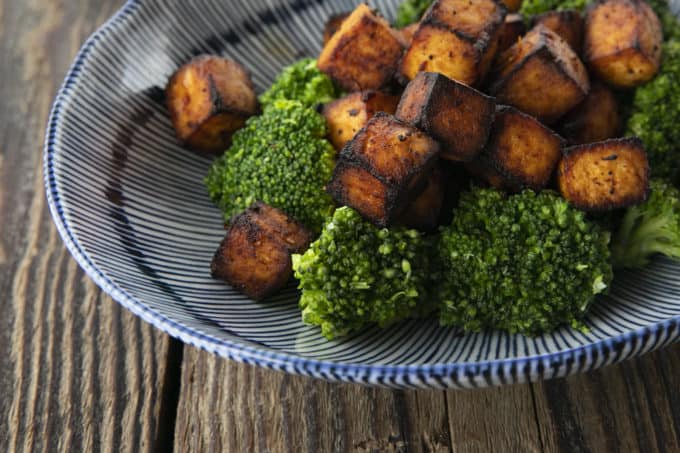 Crispy, golden brown cubes of air fryer tofu served with a drizzle of honey and broccoli on a blue and white striped oblong bowl.