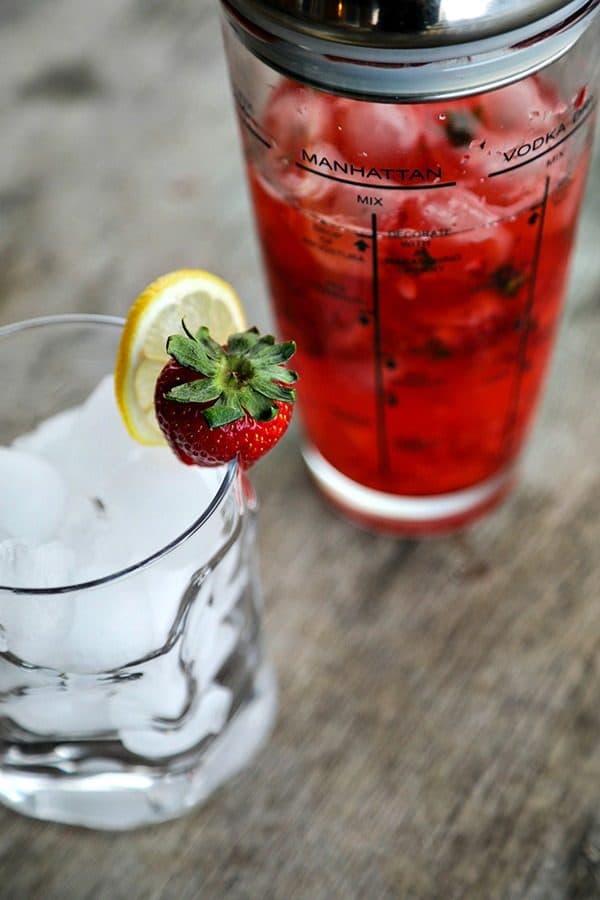 glass cocktail shaker, ice in bormioli rocco sorgente highball glass, strawberry and lemon garnish, rustic wood table