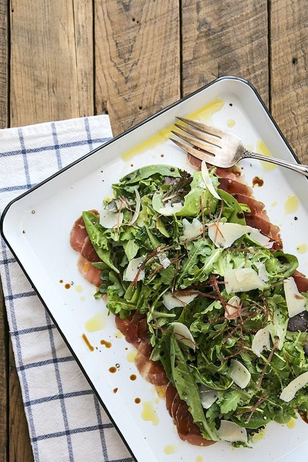 Bresaola Parmesan Salad on white quarter sheet pan with cobalt blue rim, blue and white tea towel, antique fork, rustic wood tabletop