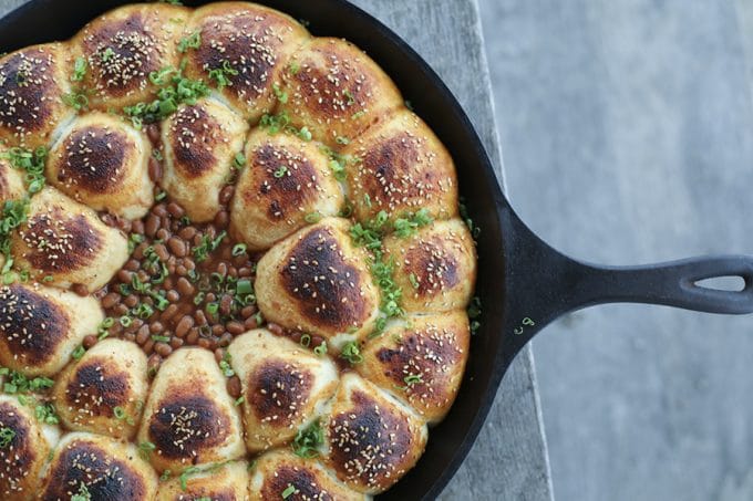 Cheeseburger Bombs Baked Bean Skillet is a whole host of comfort foods in one pan! Delicious rolls stuffed with melted Cheddar cheese and tender meatballs, baked in a cast iron skillet around barbecue baked beans. This is as good as game day food gets!