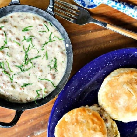 Classic Sausage Gravy in black and white enameled bowl with handles, biscuits on blue and white enamel plate, round wooden platter, fork with wooden handle, blue and white enamel metal spoon