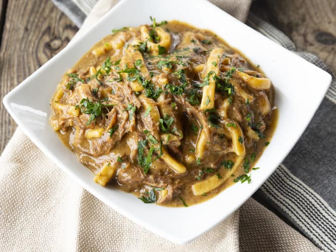 crock pot beef and noodles in a white square bowl on linens