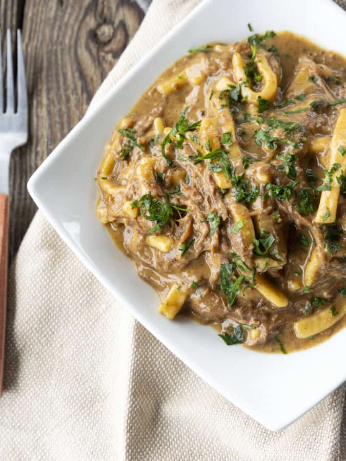 crock pot beef and noodles in a white square bowl on linens