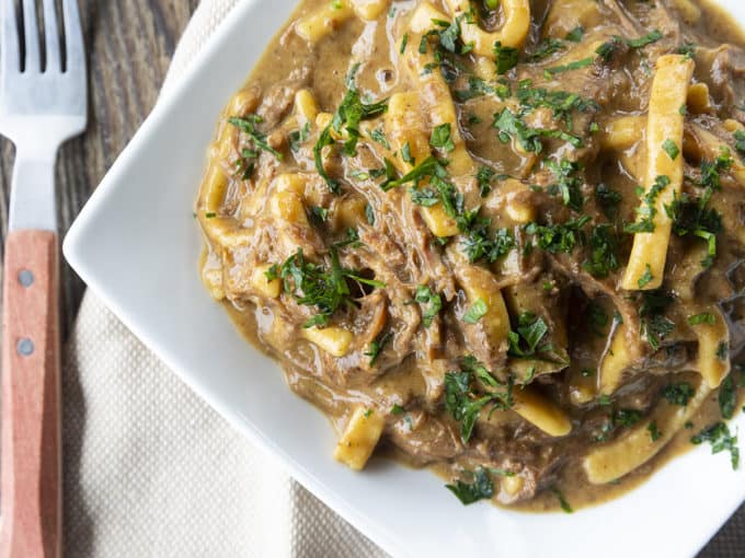 crock pot beef and noodles in a white square bowl on linens