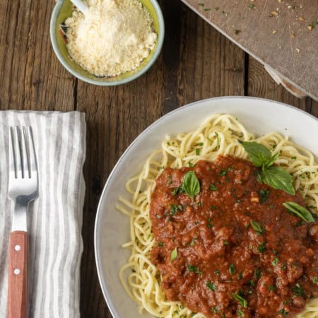 spaghetti sauce on spagetti on a plate topped by fresh herbs with a napkin and fork on the left and garlic bread on a cutting board above.