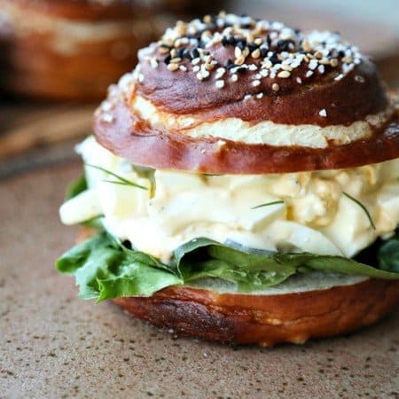 Dill Pickle Egg Salad on lettuce and pretzel roll, brown speckled pottery plate, lettuce leaves, wooden cutting board, rustic wood table, close up