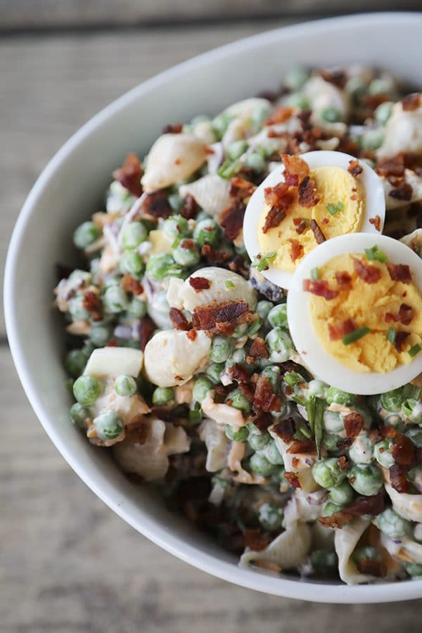 bacon pea pasta salad in white bowl with hard boiled eggs, bacon bits, rustic wood table