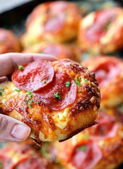 Pepperoni Pizza Bialys Recipe held by hand, melted cheese, crisp pepperoni, parsley, background of pizza on antique cookie sheet.