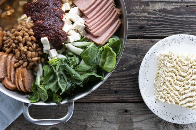 Budae Jjigae or Korean Army Stew ready to simmer. Stainless steel everyday pan filled with bok choy, sliced sausace, spam, mushrooms, tofu cubes, baked beans, and sauce. Ramen noodles on a plate next to the pan.