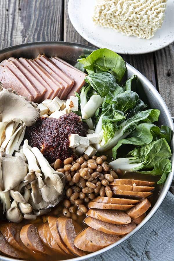 Korean Army Stew or Budae Jjigae in a stainless steel everyday pan, ready to simmer. Bok choy, sliced sausage, sliced spam, mushrooms, baked beans, gochujang seasoning, and ramen noodles on the side on a plate.