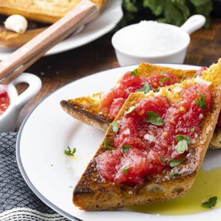 Sublime and simple Pan Con Tomate has just five ingredients; toasted rustic bread rubbed with a garlic clove, juicy fresh tomato pulp, extra-virgin olive oil, and coarse flaky sea salt. Also known as Spanish Tomato Bread, Pan Tumaca, or Pa Amb Tomàquet, this easy recipe using simple ingredients is equally at home served at a dinner party with a Spanish omelette or as part of a traditional Spanish breakfast.