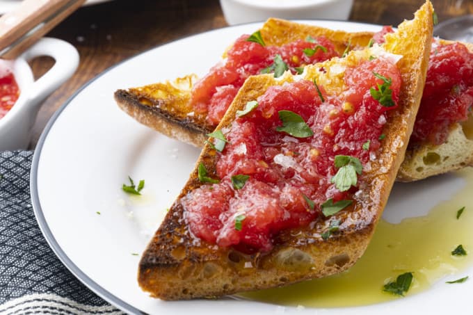 Sublime and simple Pan Con Tomate has just five ingredients; toasted rustic bread rubbed with a garlic clove, juicy fresh tomato pulp, extra-virgin olive oil, and coarse flaky sea salt. Also known as Spanish Tomato Bread, Pan Tumaca, or Pa Amb Tomàquet, this easy recipe using simple ingredients is equally at home served at a dinner party with a Spanish omelette or as part of a traditional Spanish breakfast.