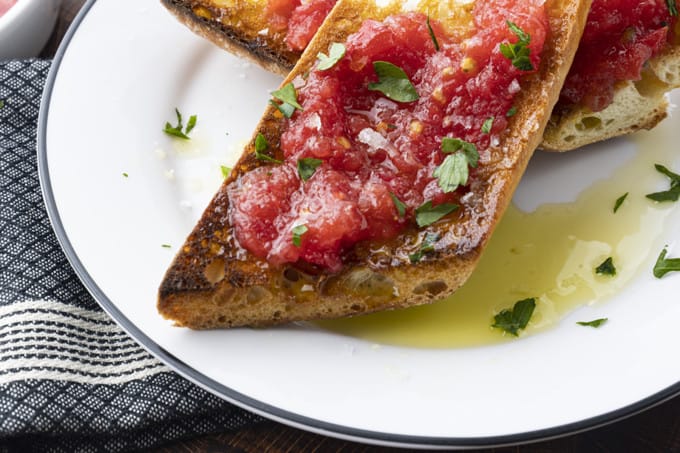 Sublime and simple Pan Con Tomate has just five ingredients; toasted rustic bread rubbed with a garlic clove, juicy fresh tomato pulp, extra-virgin olive oil, and coarse flaky sea salt. Also known as Spanish Tomato Bread, Pan Tumaca, or Pa Amb Tomàquet, this easy recipe using simple ingredients is equally at home served at a dinner party with a Spanish omelette or as part of a traditional Spanish breakfast.