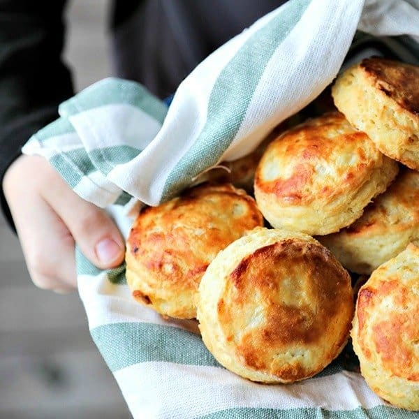 perfect flaky buttermilk biscuits in green and white towel lined bowl, held by boy in black long sleeved shirt, wooden porch