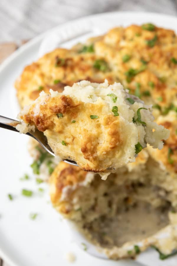 small white casserole dish filled with sausage gravy and topped with buttermilk biscuit crust on top of a plate with a spoon in it.