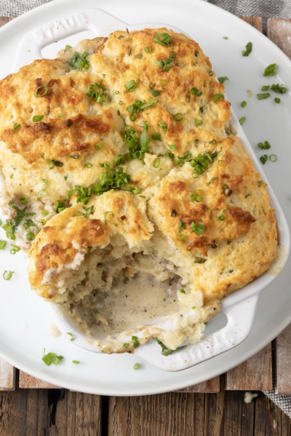 small white casserole dish filled with sausage gravy and topped with buttermilk biscuit crust on top of a plate with a scoop out of it