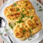 small white casserole dish filled with sausage gravy and topped with buttermilk biscuit crust on top of a plate with a spoon in it with descriptive text overlay.