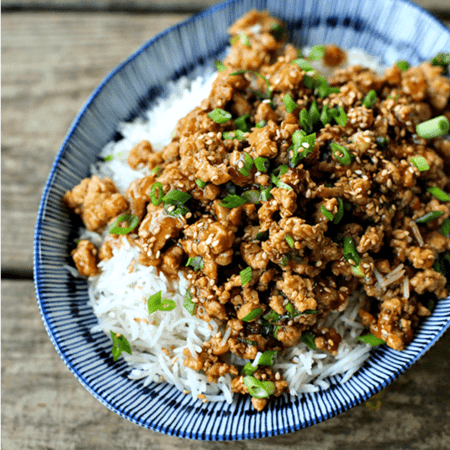 Cheater Sesame Chicken ground chicken in sweet and spicy sauce, sesame seeds, sliced green onions, long grain white rice, asian bamboo pattern blue and white bowl on wooden bench